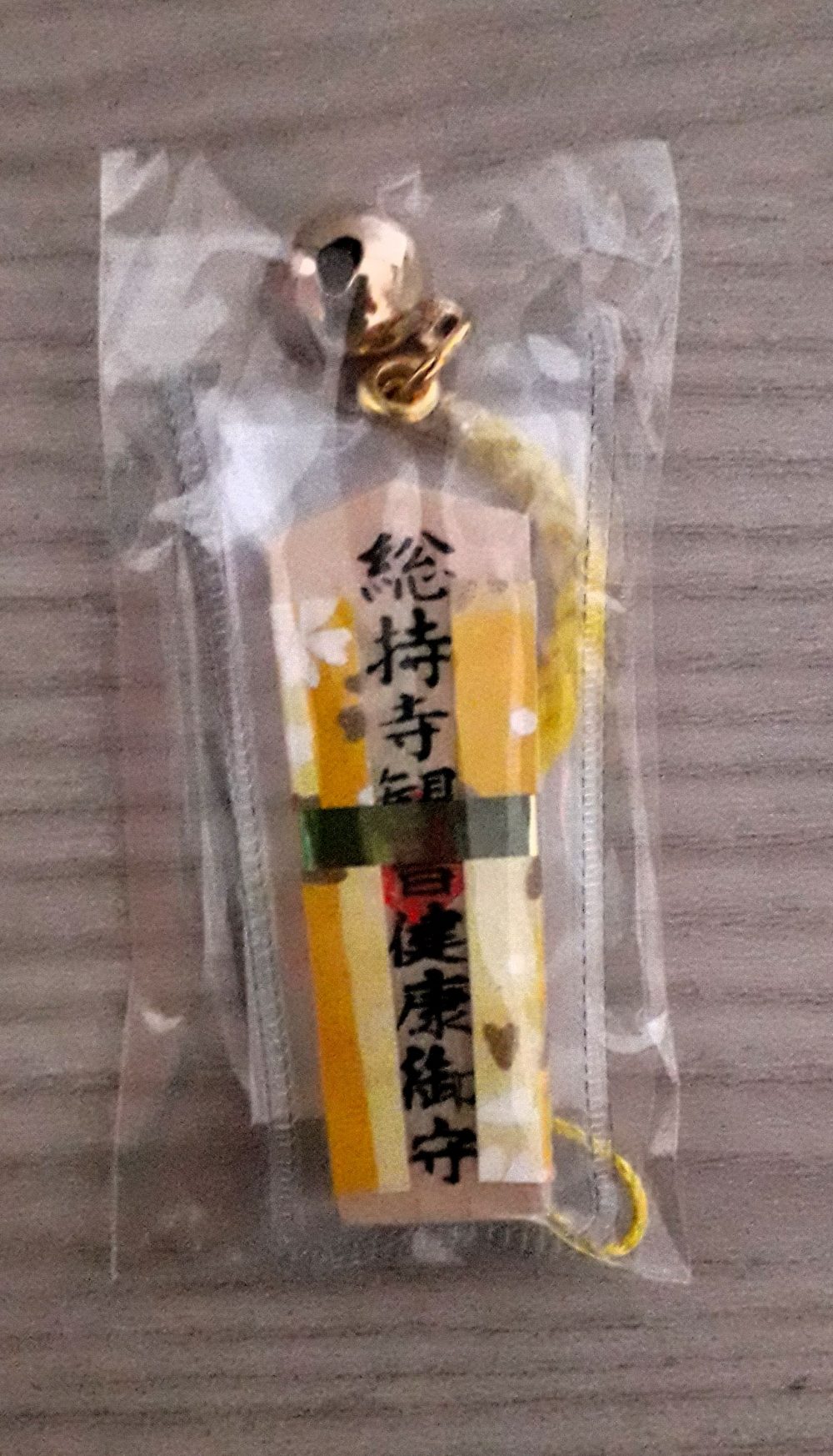 Omamori a ciondolo in legno, rivestito con stoffa di colore giallo, per augurare o augurarsi una buona salute.