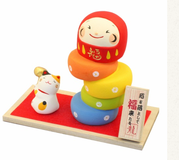 Yakuotoshi Daruma, questa statuina rappresenta un gatto calico che sta giocando ad un tipico gioco giapponese, ovvero "Daruma Otoshi". A "Daruma Otoshi" si gioca posizionando una bambola daruma sopra ad una "torre" di 5 cerchi, e con un piccolo martello bisogna colpire ognuno dei pezzi dall'alto verso il basso, senza far cadere gli altri.